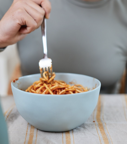 Person eating spaghetti with a fork from a light blue bowl on a patterned tablecloth.